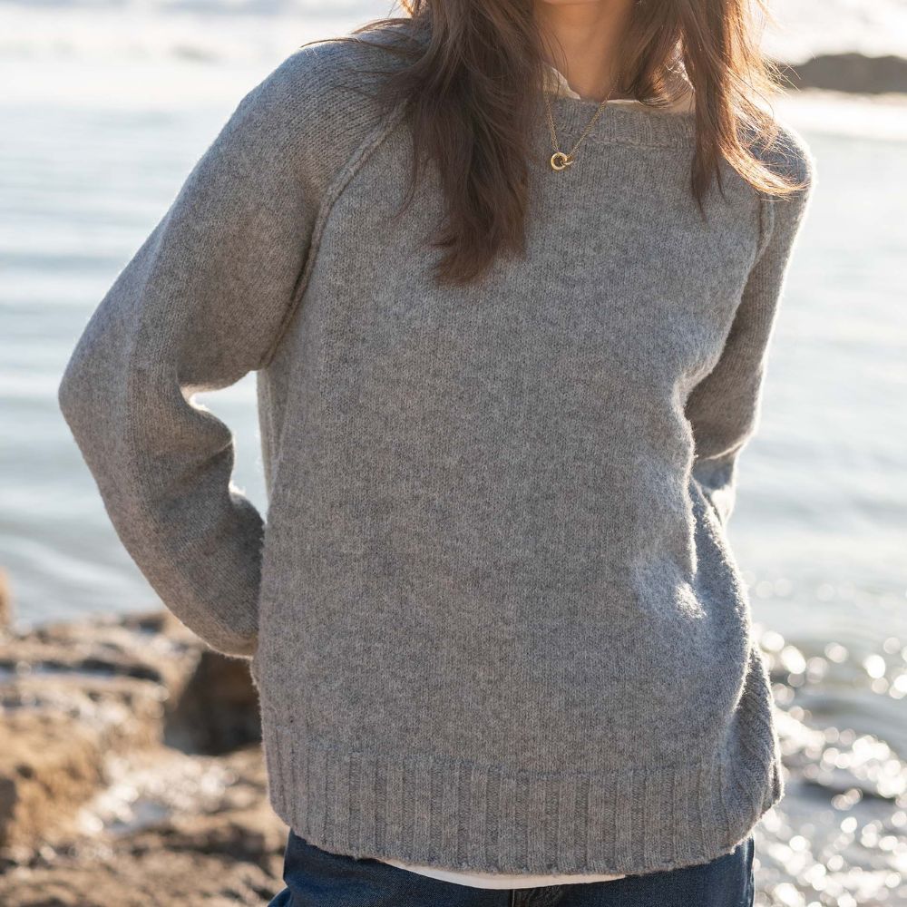 A woman wearing a light gray knit sweater with raglan sleeves, standing outdoors near the water.