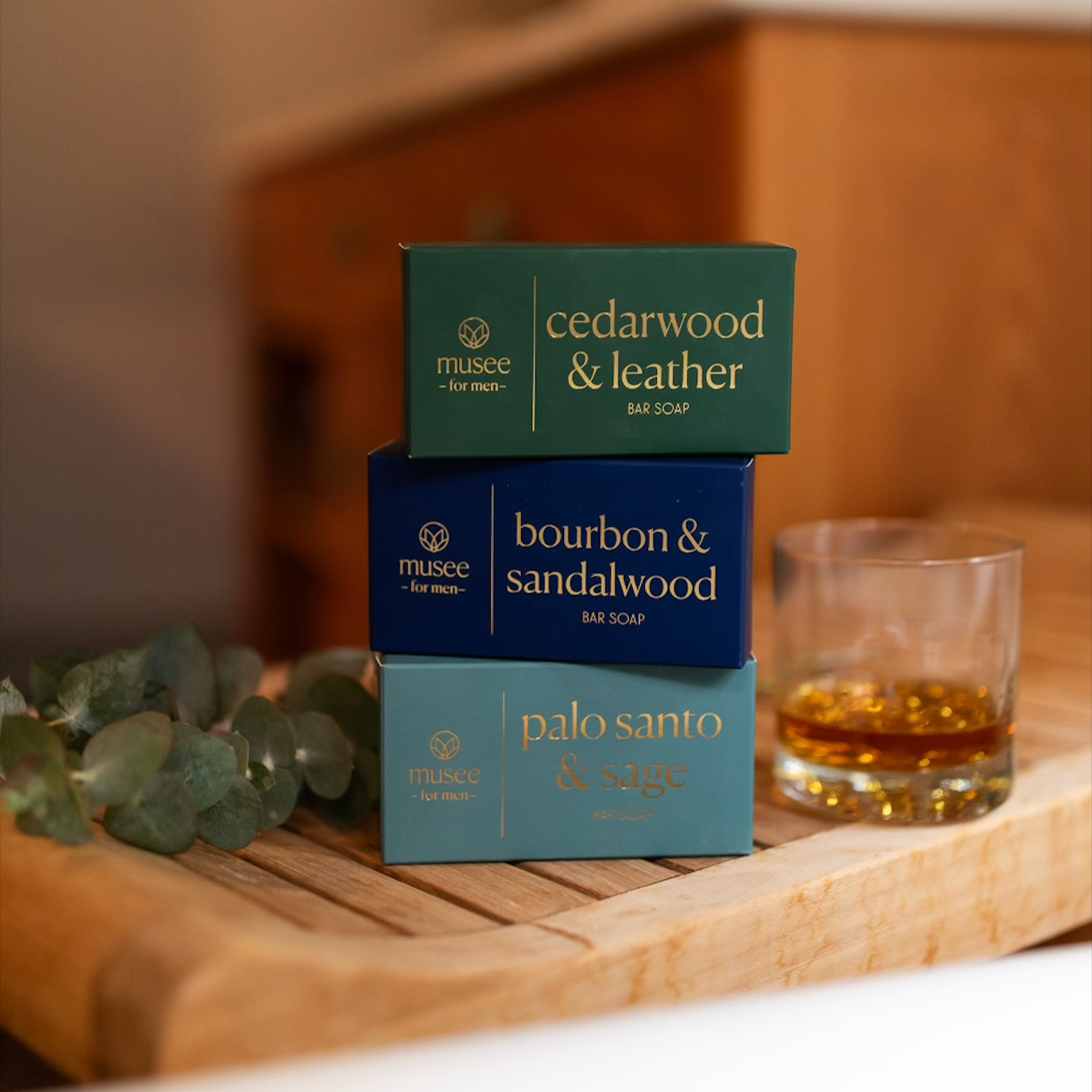 Three bar soaps vertical on a wooden bath surface next to a glass of bourbon