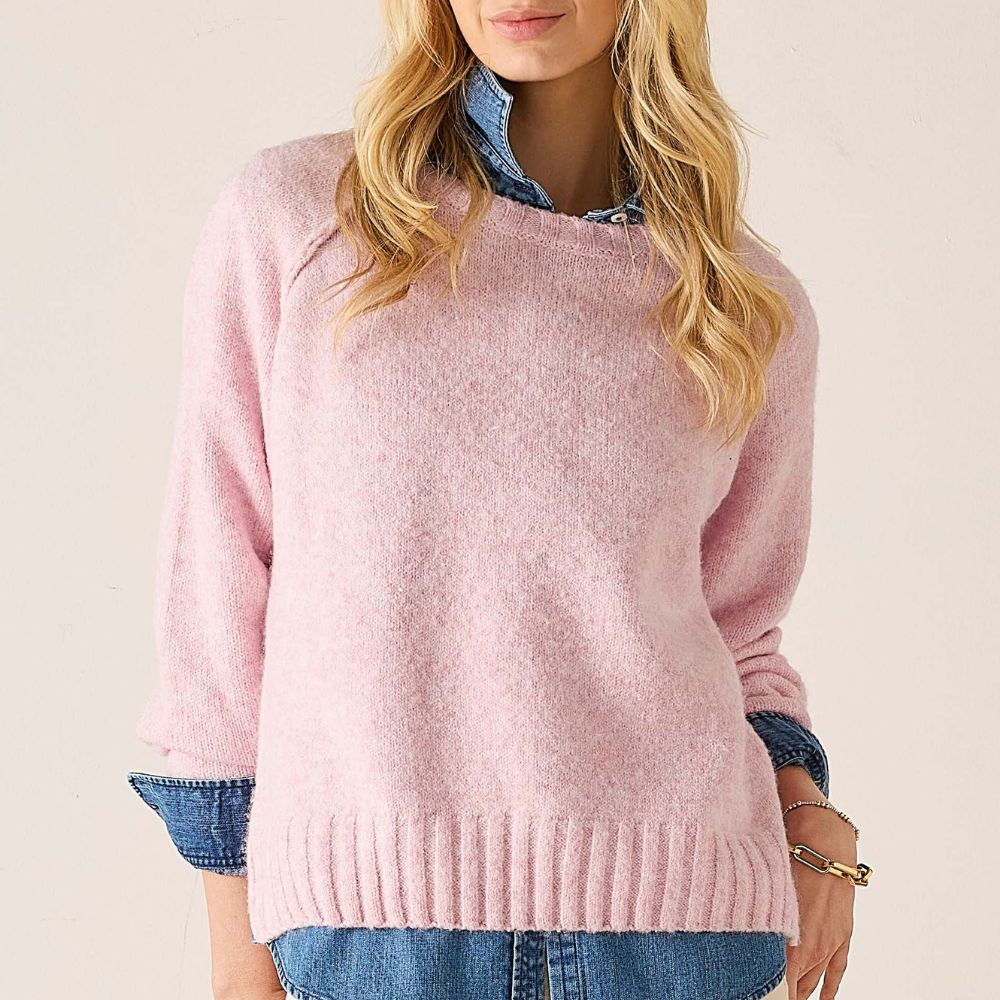 A woman wearing a soft pink knit sweater with a crew neck and ribbed hem, layered over a blue denim shirt and paired with blue jeans, against a plain light beige background.