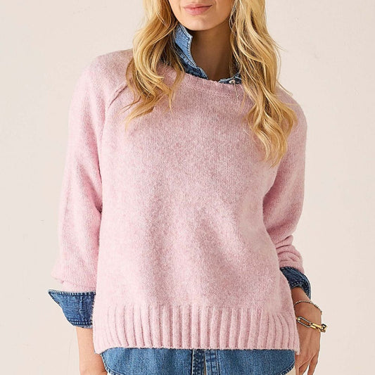 A woman wearing a soft pink knit sweater with a crew neck and ribbed hem, layered over a blue denim shirt and paired with blue jeans, against a plain light beige background.