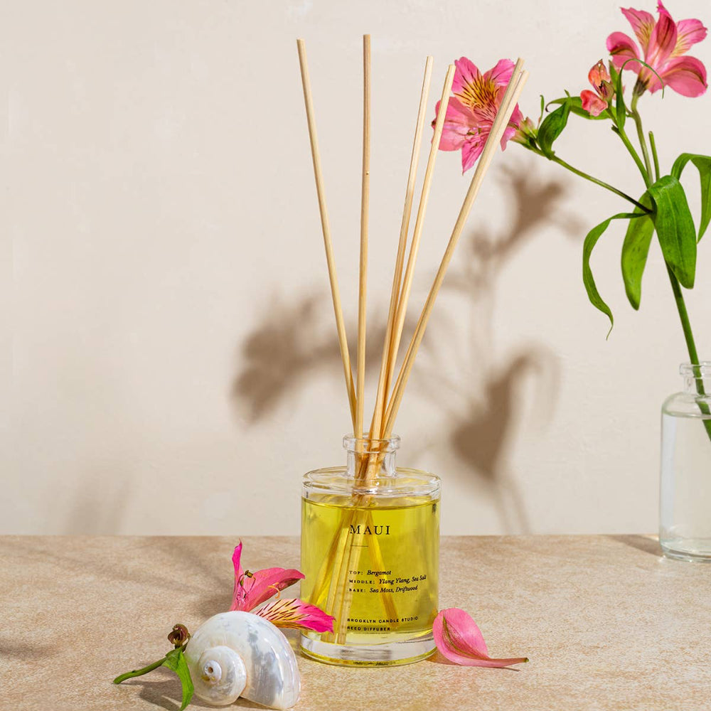 Maui reed diffuser with rattan reeds on a wood surface with pink flowers and a shell