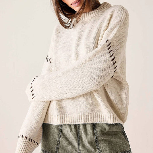 A woman wearing a cream-colored knit sweater with black geometric cutout details along the sleeves, paired with olive green denim shorts, against a plain light beige background.