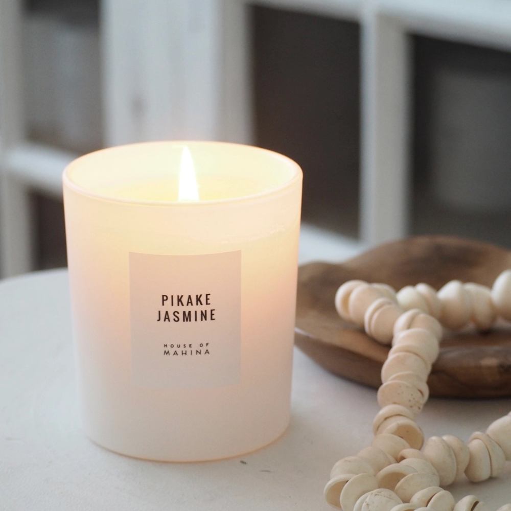 Lit Pikake Jasmine candle on counter top with wood bowl and strand of beads in the background