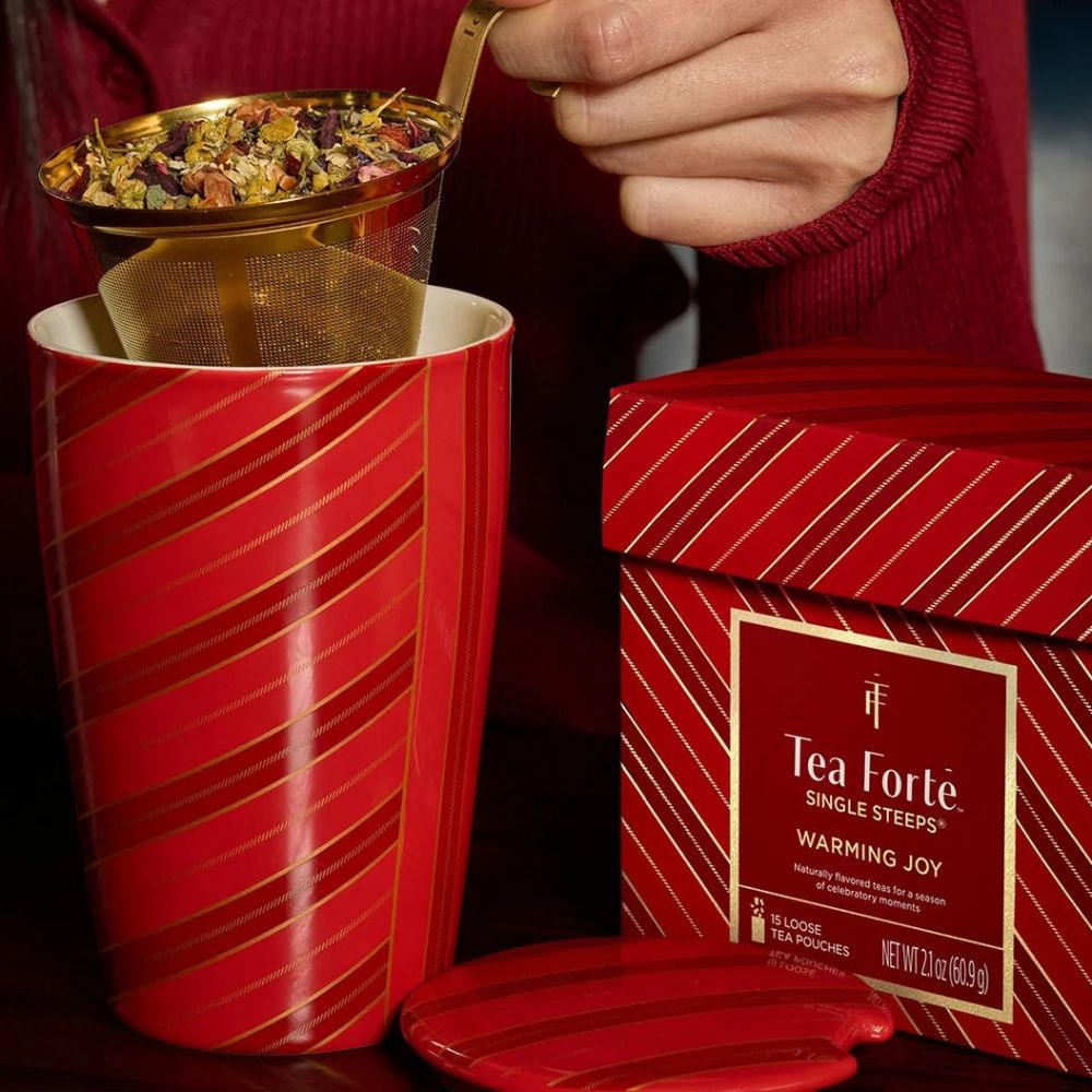 Hand spooning loose tea into a gold infuser basket over the Warming Joy Kati cup, Single Steeps box beside it